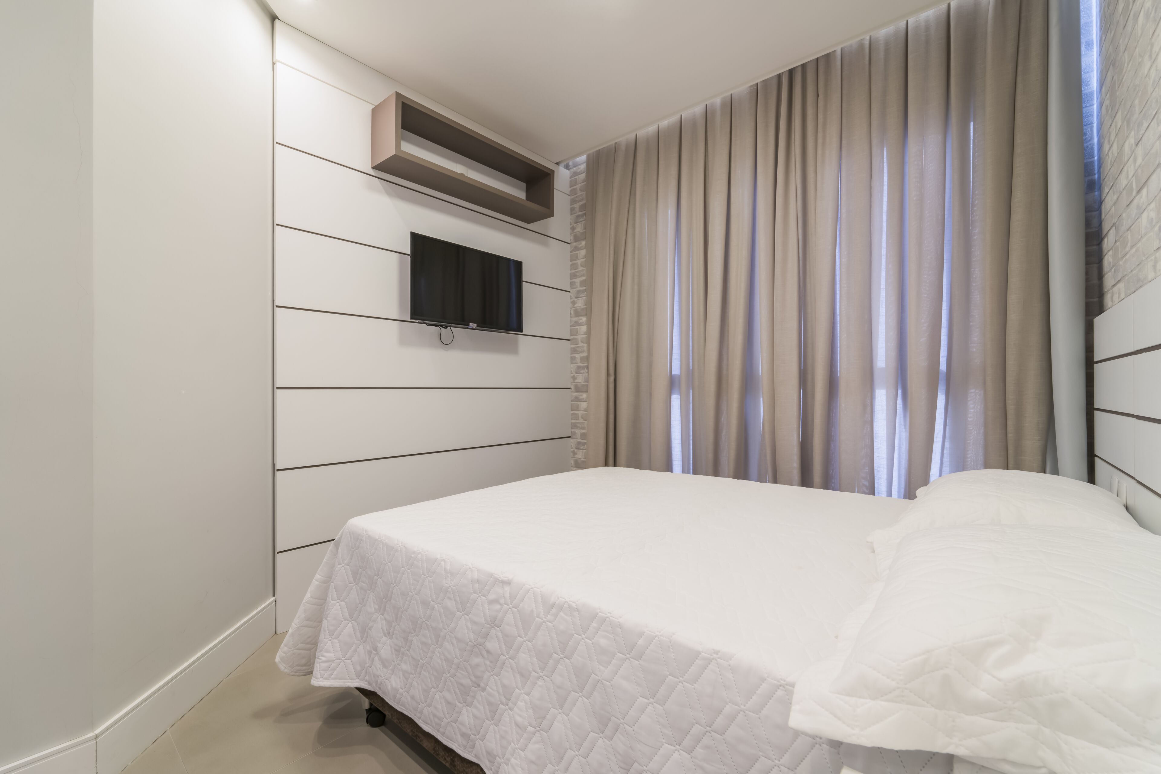 A bedroom with a bed, a mounted TV, and a window with curtains.