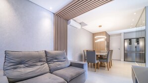 Een moderne woonkamer met een grijze bank, een eethoek met een houten tafel en stoelen, en een keuken met een roestvrijstalen koelkast.