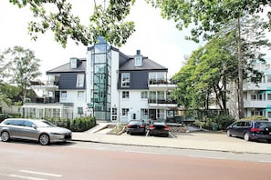 A modern building with a glass facade, a parking area, and a street with cars. - Modernes Strandhus Appartement 109 (Timmendorfer Strand)