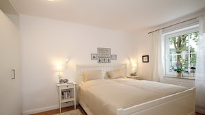 Un dormitorio con una cama grande, mesitas de noche, una lámpara de noche, un cuadro enmarcado y una ventana con cortinas.