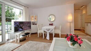 Una sala de estar moderna con televisor, muebles blancos, una mesa de comedor y un jarrón de flores.