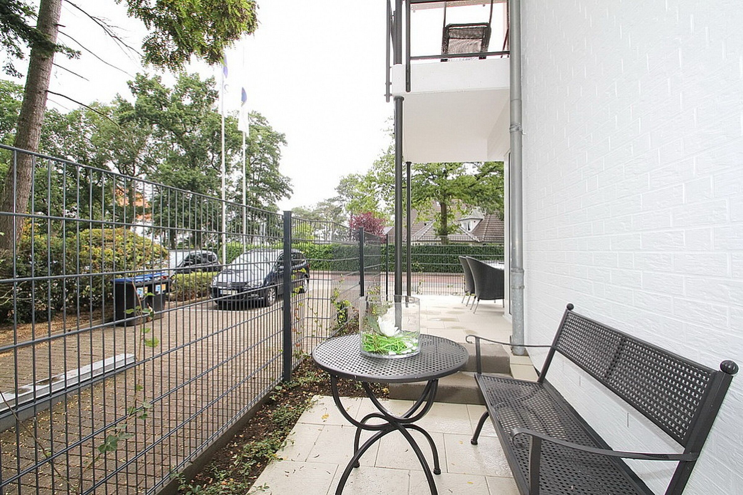 Un balcon doté d’une table et d’un banc en métal, entouré d’une clôture métallique, offrant une vue sur une rue et des voitures garées.