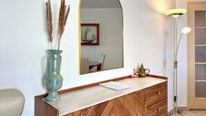 Une pièce avec une coiffeuse en bois, un miroir, un vase contenant des plumes d’herbe des pampas, une lampe et un tableau encadré au mur.