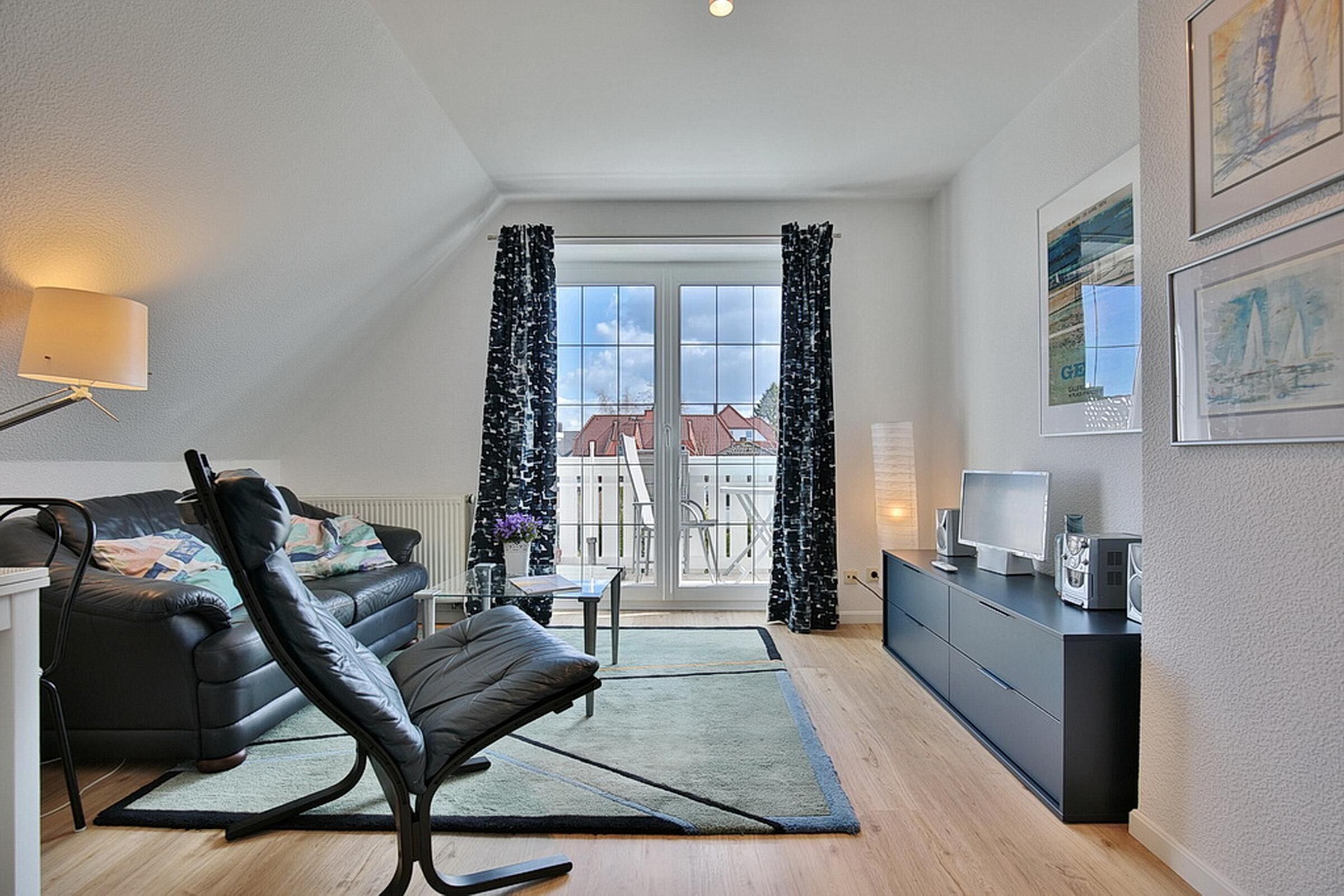 A modern living room with a leather sofa, a black rocking chair, a small table, and a TV on a black stand. There is a large window with curtains, a radiator, and a view of buildings outside.