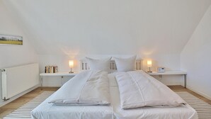 Een slaapkamer met een schuine plafond, twee bedden met witte beddengoed, een nachtkastje met een lamp, een kleine tafel met boeken, en een radiator.