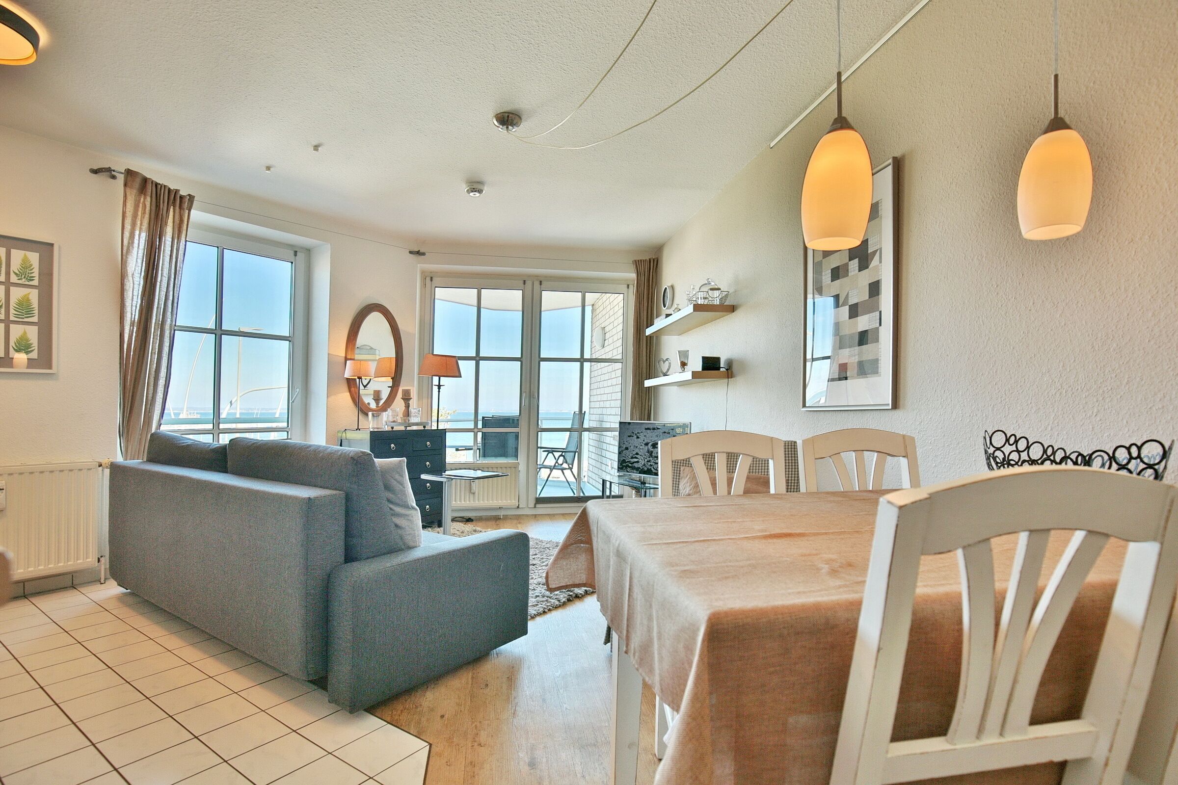 A modern living room with a sofa, dining table, and chairs, large windows with curtains, and a view of the sea.