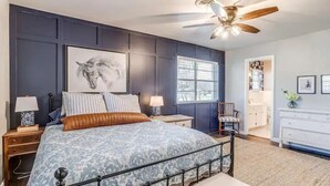 Une chambre à coucher comprenant un lit, des tables de chevet, un ventilateur de plafond, une fenêtre et une porte donnant sur une autre pièce.