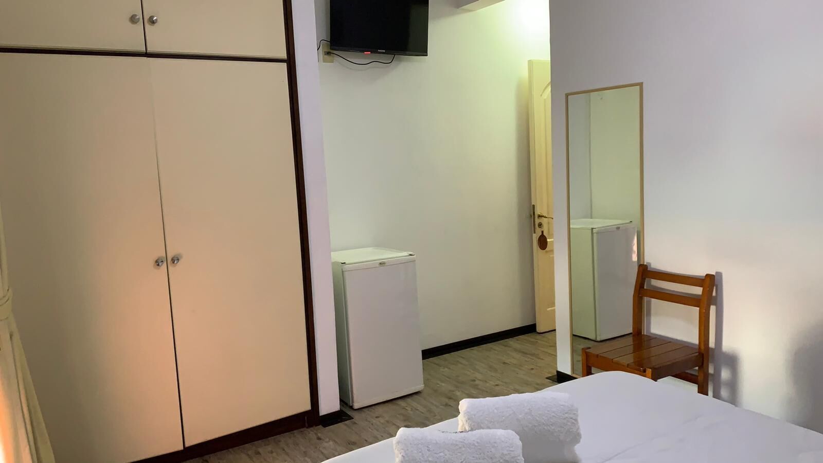 Ein Hotelzimmer mit einem Bett, einem Holzstuhl, einem Kleiderschrank, einer kleinen weißen Kommode und einem an der Wand befestigten Fernseher.