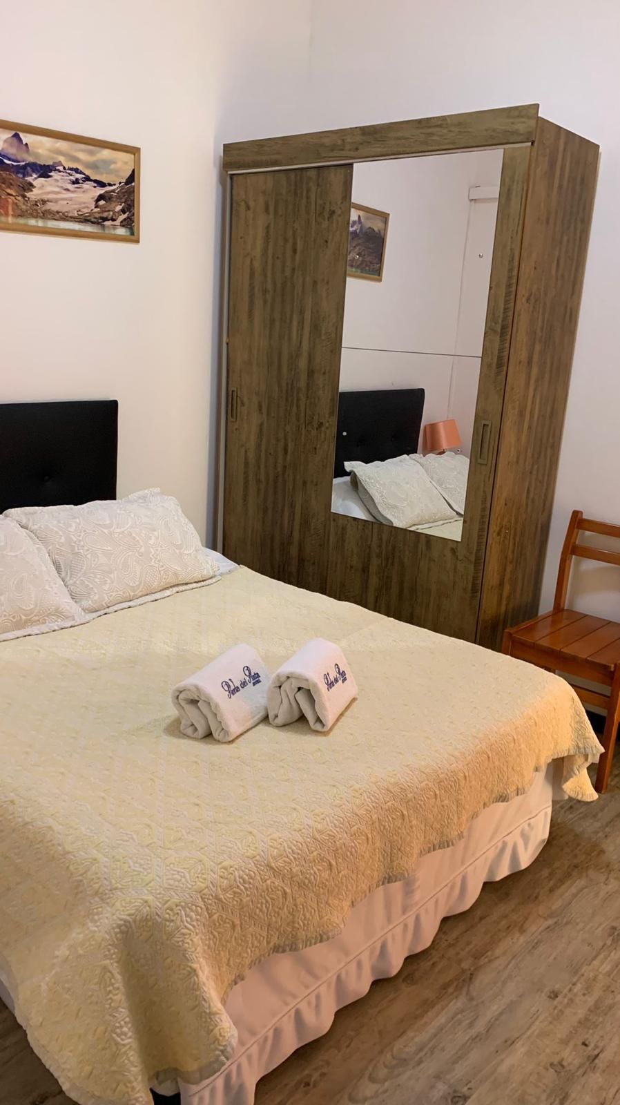 Una cama con una colcha blanca, dos toallas enrolladas, un armario de madera y un cuadro en la pared.