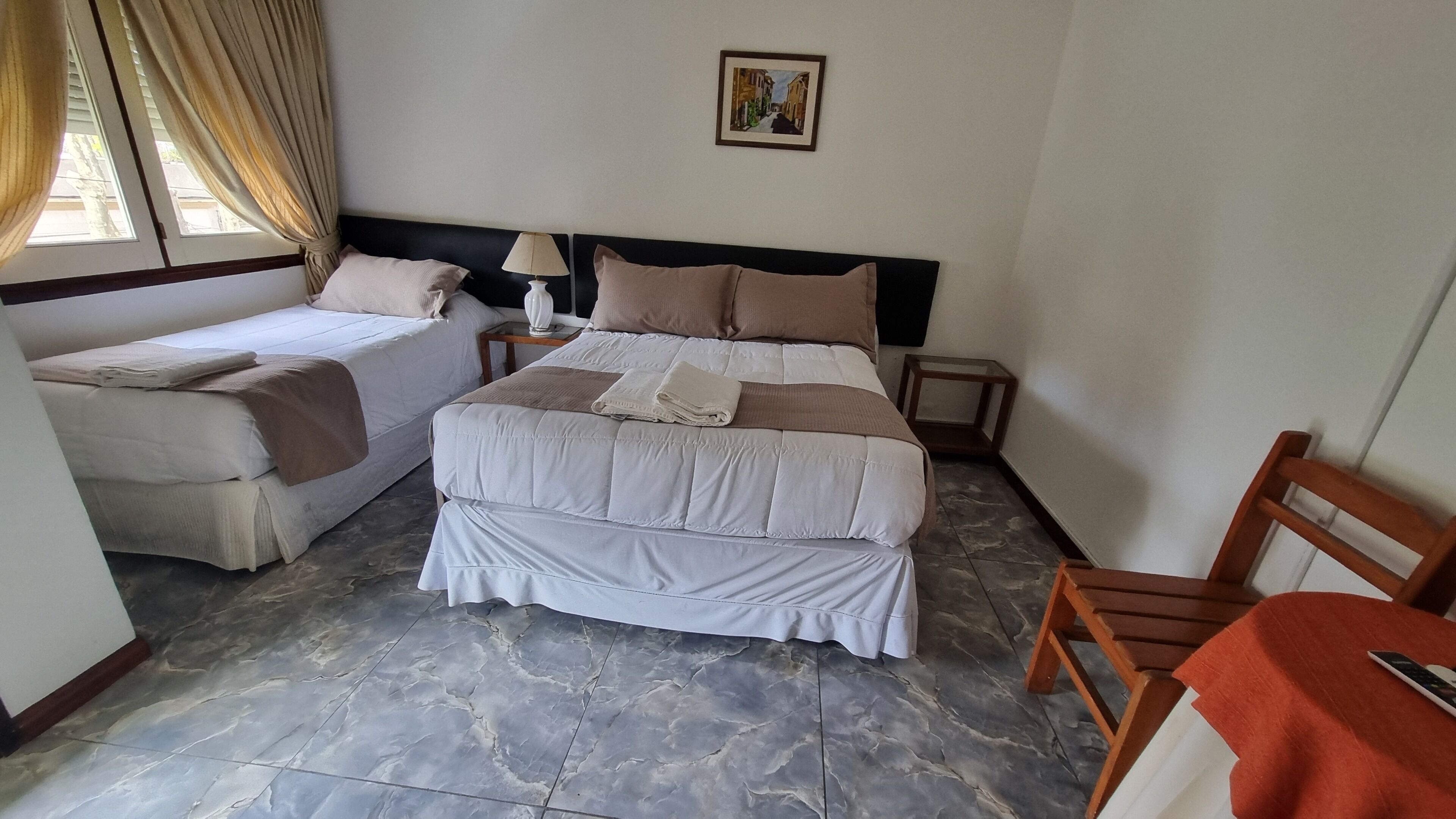 Une chambre d’hôtel avec deux lits, une table de chevet avec une lampe, un tableau encadré au mur et une chaise avec une petite table.