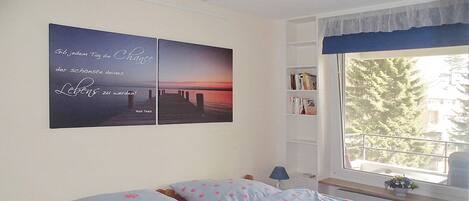 Un dormitorio con cama, mesita de noche, estantería y ventana con vistas a árboles.