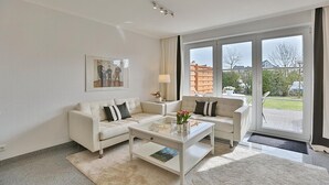 Un salon moderne avec un canapé blanc, une table basse en verre et une grande fenêtre donnant sur l’extérieur.