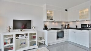 Une cuisine moderne avec des meubles blancs, un four encastré, un four à micro-ondes et un téléviseur posé sur un meuble console blanc.
