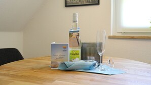 Ett bord med en flaska, ett glas, en vikt servett och ett kort på ett träbord i ett rum med ett fönster och en inramad tavla på väggen.