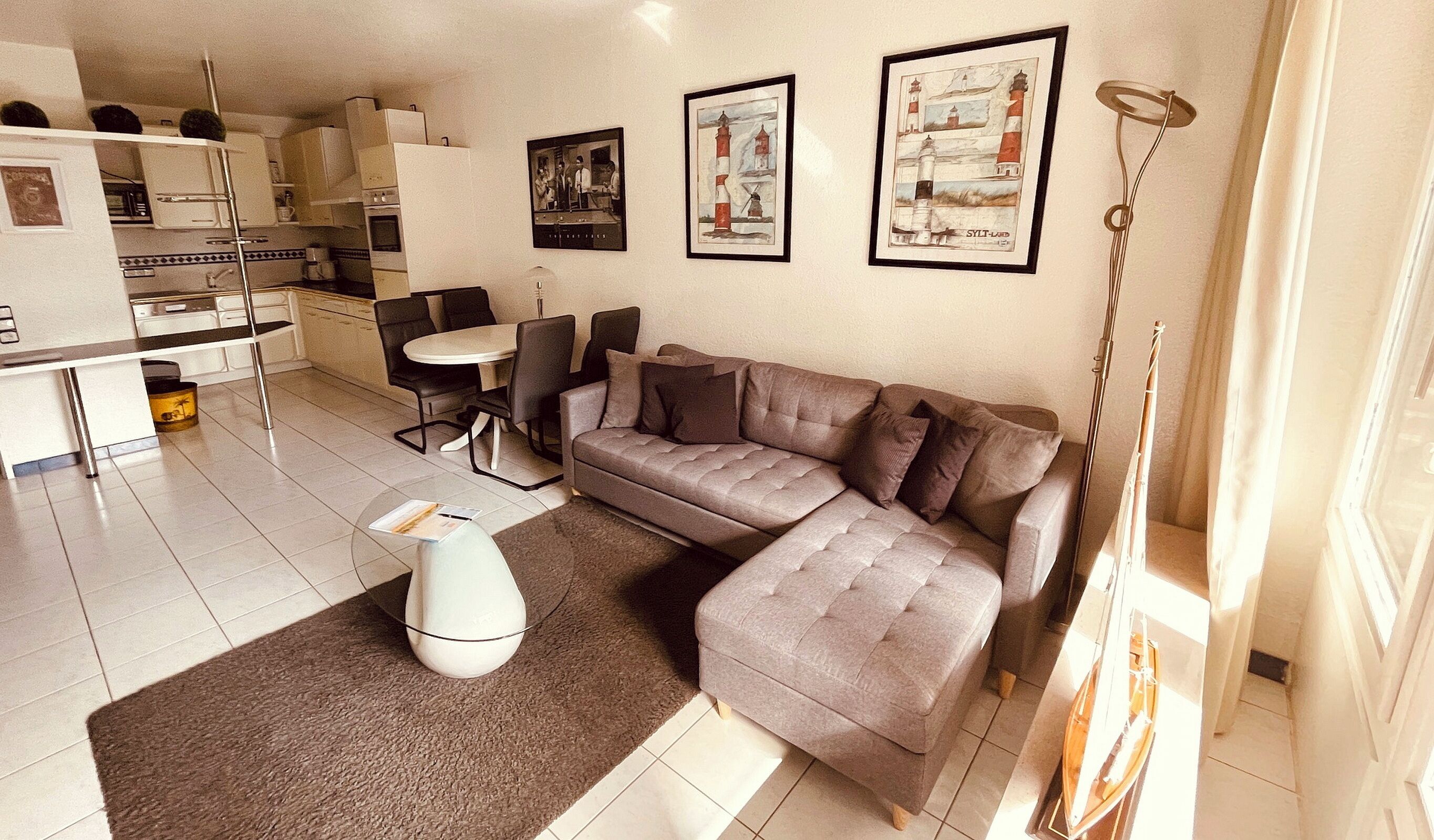 Una sala moderna con un sofá seccional gris, una mesita redonda pequeña y una mesa de centro blanca. Hay cuadros enmarcados en la pared y una zona de cocina al fondo.