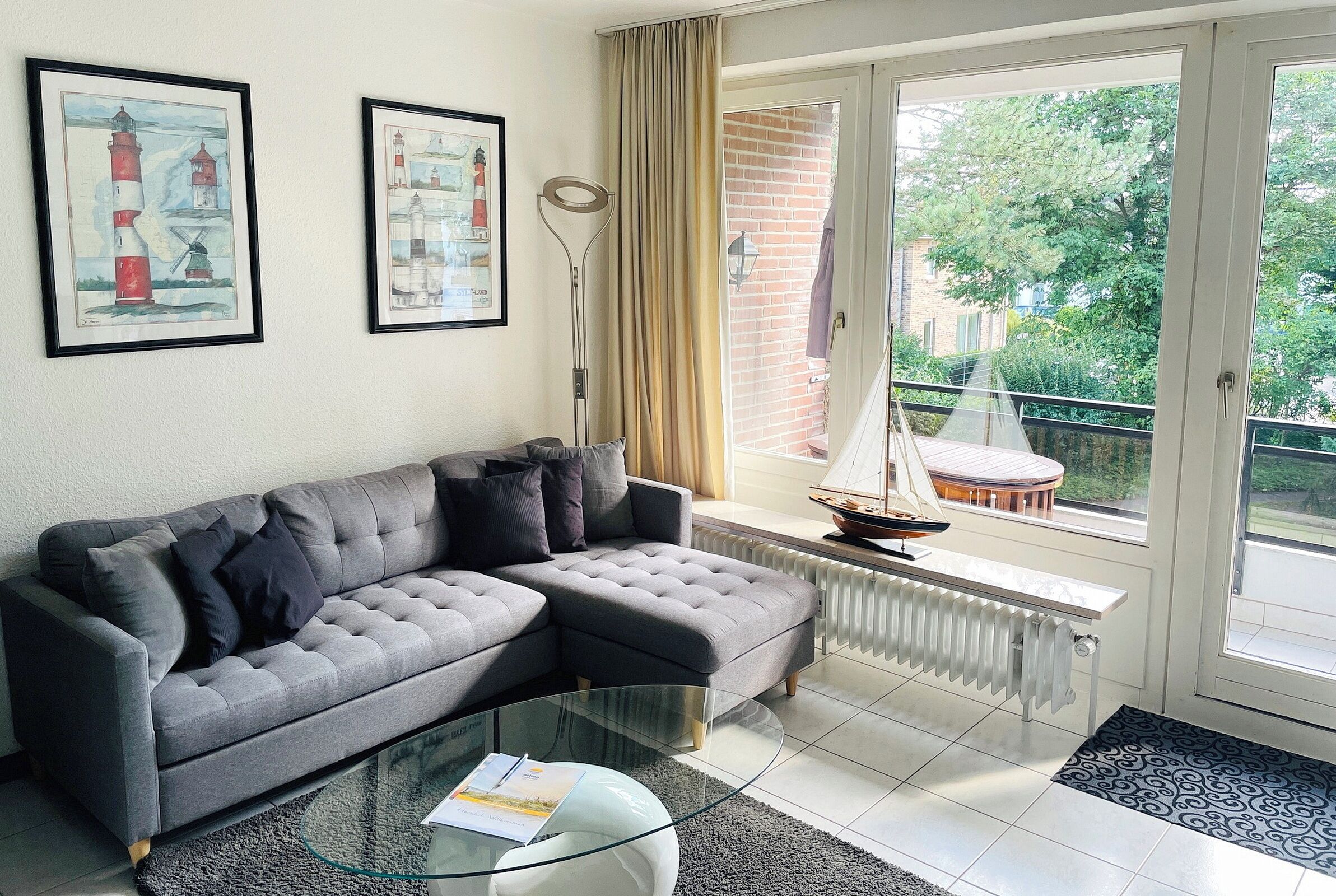 A living room with a grey sofa, a glass coffee table, and a view of a balcony with a sailboat.