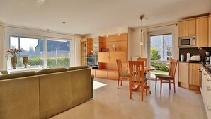 A modern kitchen with wooden cabinets, a dining area with a table and chairs, and a sofa in the living room. - Wohldkamp 4 Appartement 4 (Timmendorfer Strand)