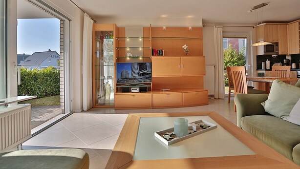 A modern living room with a glass coffee table, a sofa, and a kitchen area with wooden cabinets and a dining table. - Wohldkamp 4 Appartement 4 (Timmendorfer Strand)