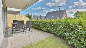 A patio with two wicker chairs and a table, a yellow wall, a hedge, and a house with a sloped roof in the background. - Wohldkamp 4 Appartement 4 (Timmendorfer Strand)