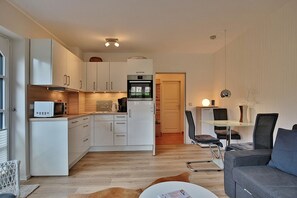 A compact kitchen with white cabinets, a microwave, and a refrigerator. There is a dining area with a glass table and chairs, and a sofa. - Am Rosenhain 23 Appartement 1 (Timmendorfer Strand)