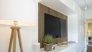A modern interior with a large flat-screen TV mounted on a wall with vertical wooden slats, a floor lamp, a wicker basket with plants, and a small bell. - Am Rosenhain Appartement 3 Hygge (Timmendorfer Strand)