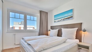 Quarto moderno com uma cama grande, uma mesinha de cabeceira, uma janela com vista para edifícios e um quadro com tema praiano na parede.
