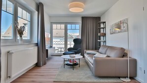 Uma sala de estar moderna com sofá, poltrona e uma pequena mesa. Há uma janela grande com cortinas, um radiador e um lustre.