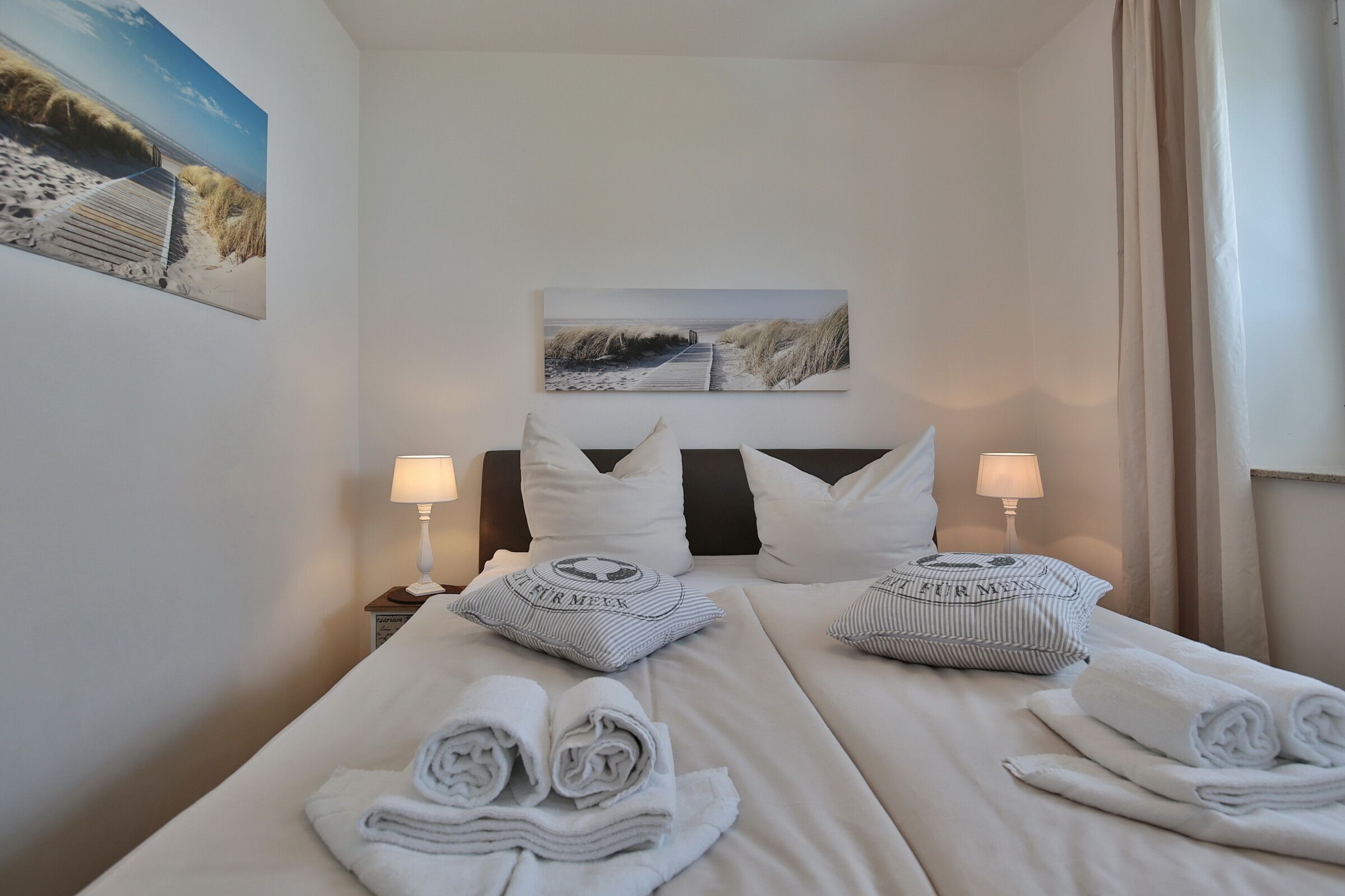 Ett hotellrum med två sängar, vita sängkläder, två kuddar med text på, två sänglampor och en tavla med ett strandmotiv på väggen.