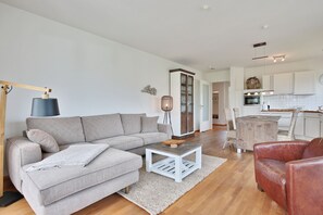 A modern living room with a grey sectional sofa, a coffee table, and a red leather chair. A kitchen with white cabinets and a wooden island is visible in the background.