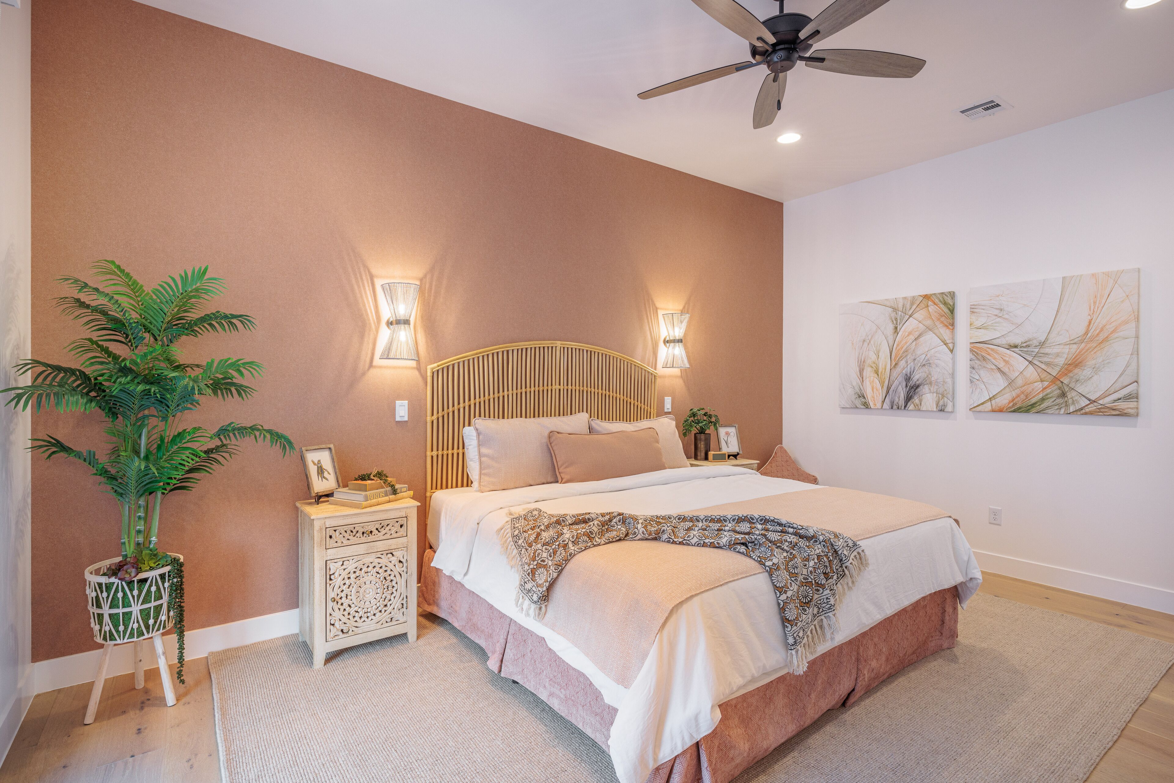 A bedroom with a bed, bedside tables, a ceiling fan, wall-mounted lights, a potted plant, and artwork.