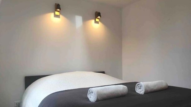 Een net opgemaakt bed met een zwart-wit dekbed, twee opgerolde handdoeken op het bed, en twee wandlampen.