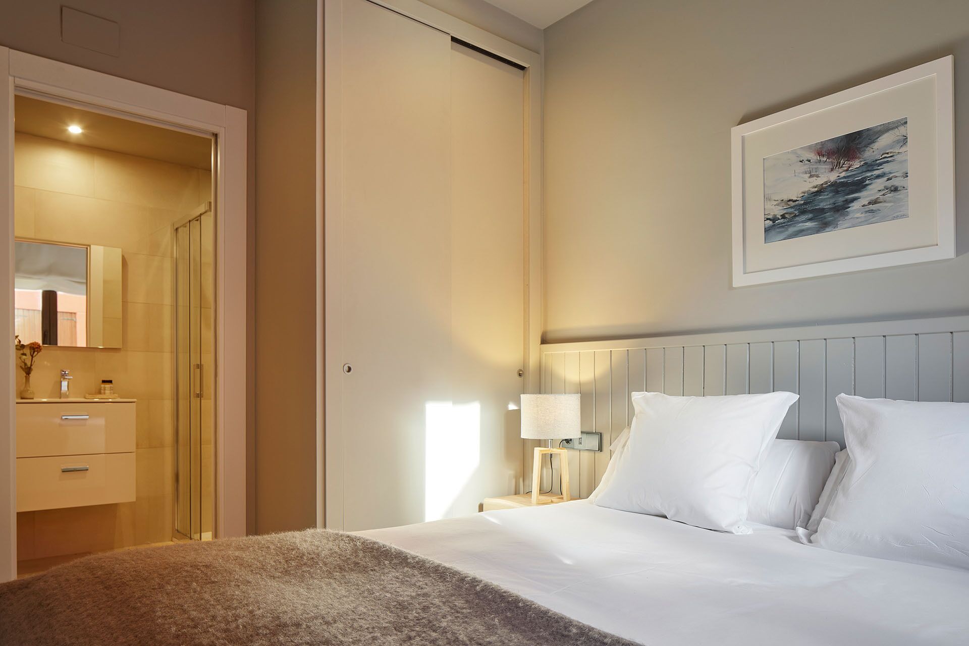 A hotel room with a bed, a nightstand with a lamp, a framed picture on the wall, and a bathroom visible through an open door.