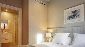 Habitación de hotel con cama, mesita de noche con lámpara, un cuadro enmarcado en la pared y un baño visible a través de una puerta abierta.