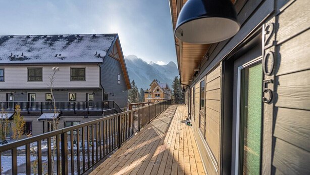 A wooden deck with a railing, a hanging lamp, and a view of snow-covered buildings and mountains in the background.