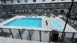 A rectangular swimming pool with a smaller adjacent hot tub, surrounded by a black metal fence, with a multi-story building in the background.