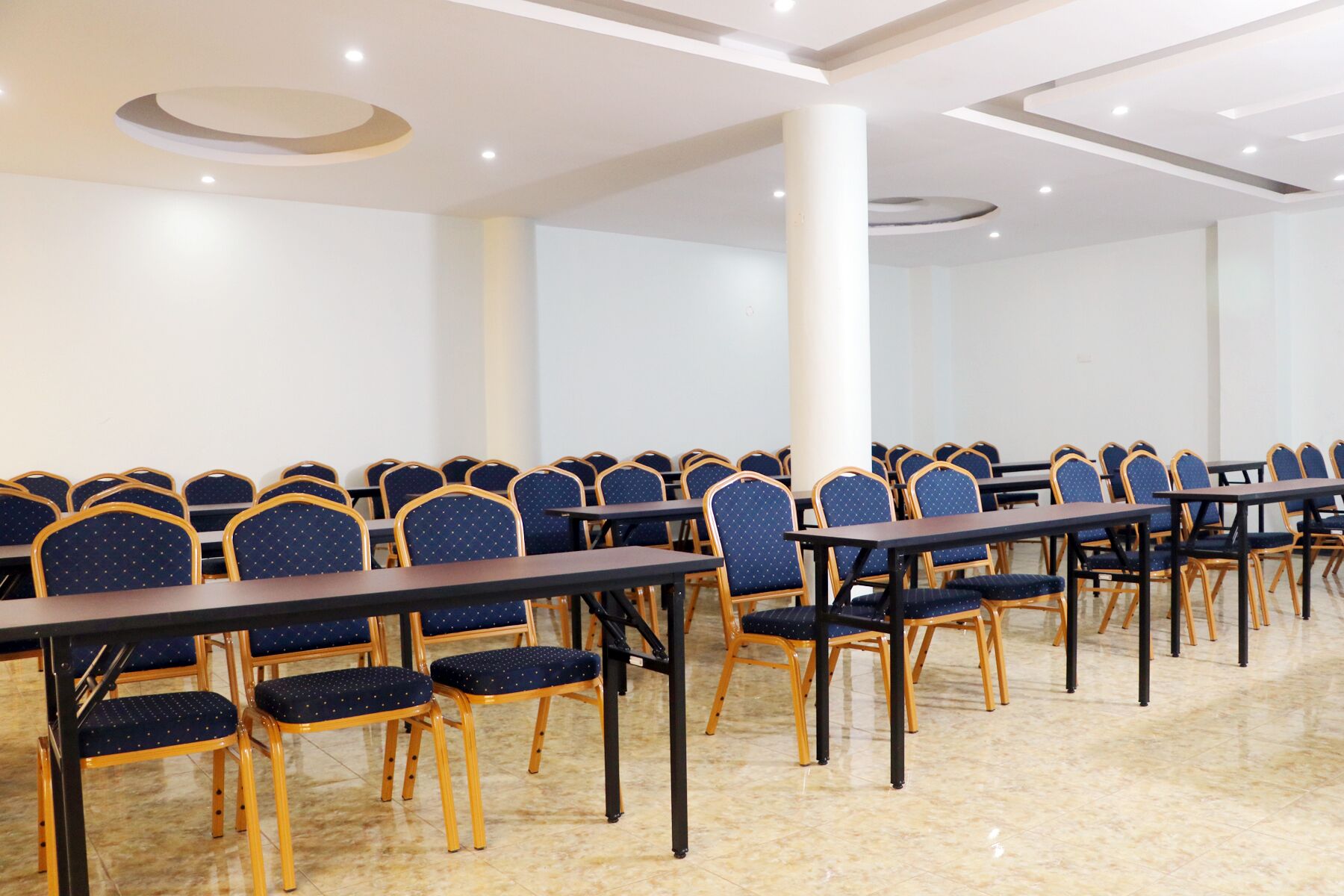 A conference room with rows of tables and chairs, white walls, and a tiled floor.