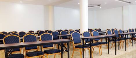 A conference room with rows of tables and chairs, white walls, and a tiled floor.