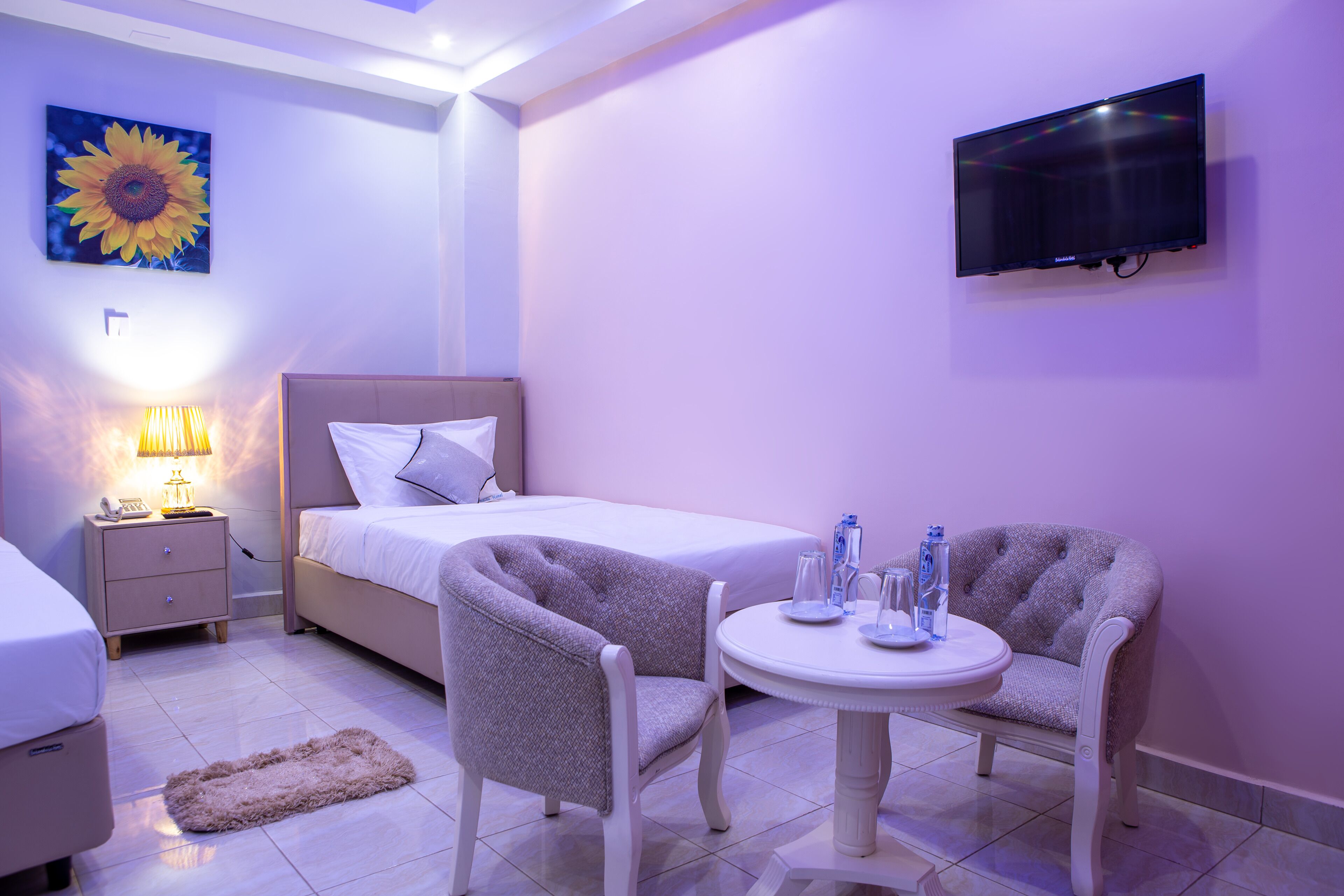 A hotel room with a bed, bedside table, lamp, TV, two armchairs, and a small table with glasses and water bottles.