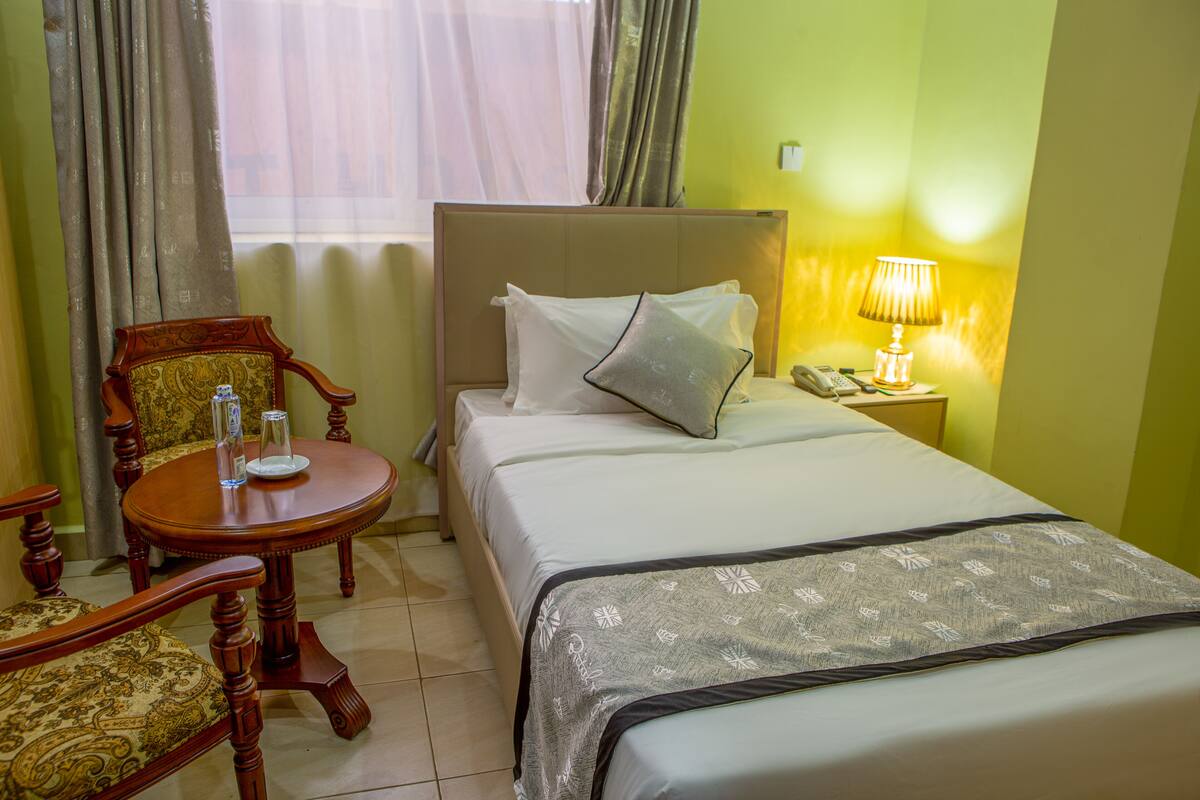 A hotel room with a bed, a wooden chair, a small table with a glass of water, and a lamp.