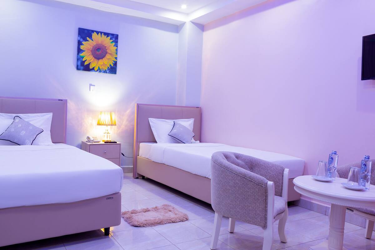 A hotel room with two beds, a nightstand with a lamp, a chair, and a small table with water bottles.