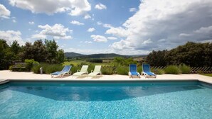 A swimming pool with clear blue water, surrounded by lounge chairs and a view of a green landscape with trees and hills in the background. - Beautiful manor with swimming pool in front of the Provençal Colorado (Rustrel)