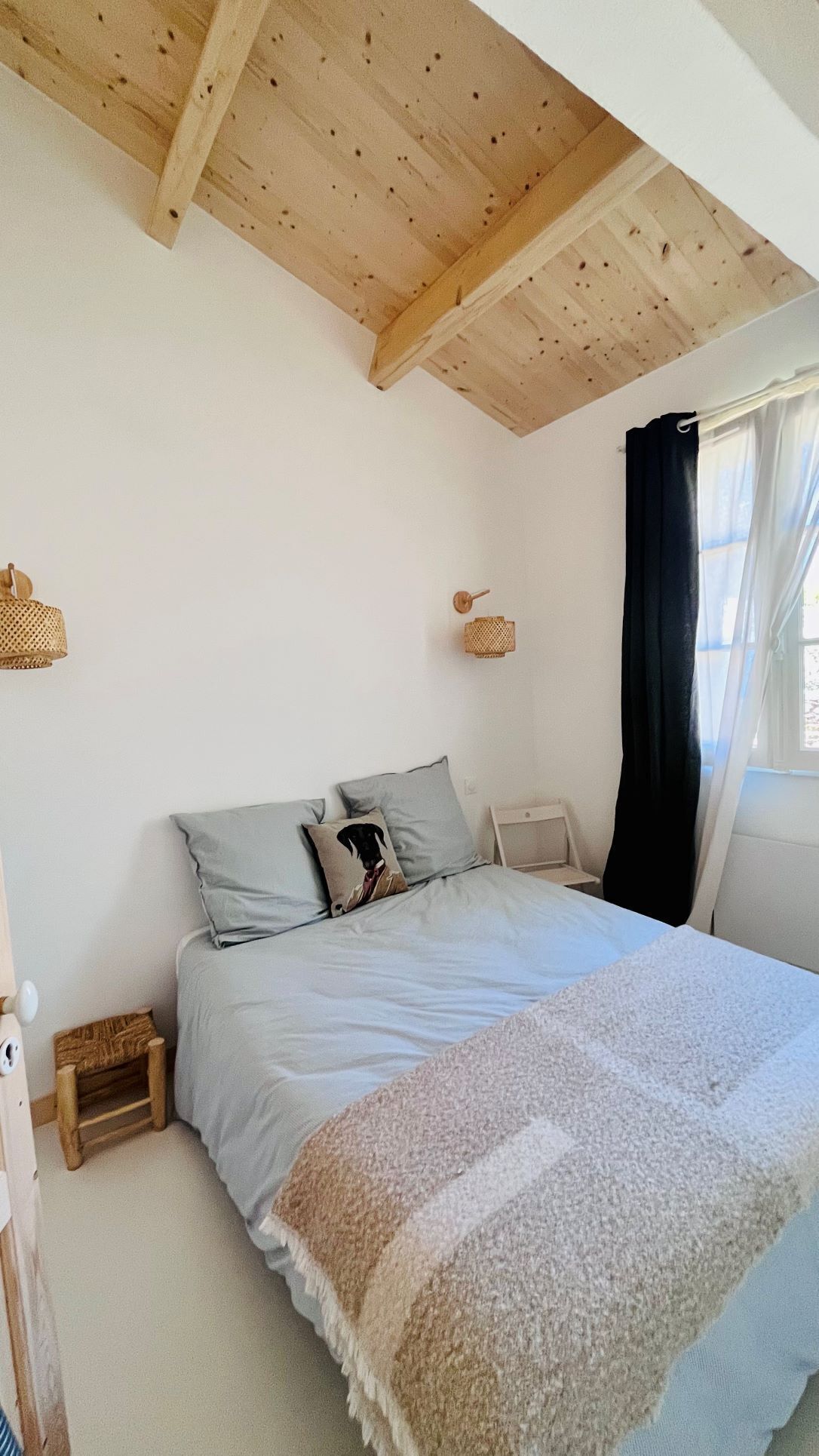 A bedroom with a wooden ceiling, a bed with grey bedding, a small bedside table, a wall-mounted lamp, and a window with a black curtain.