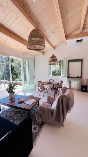Una habitación amplia con techo de madera, un comedor con una mesa redonda y sillas, un sofá y un gran ventanal con vistas al verdor.