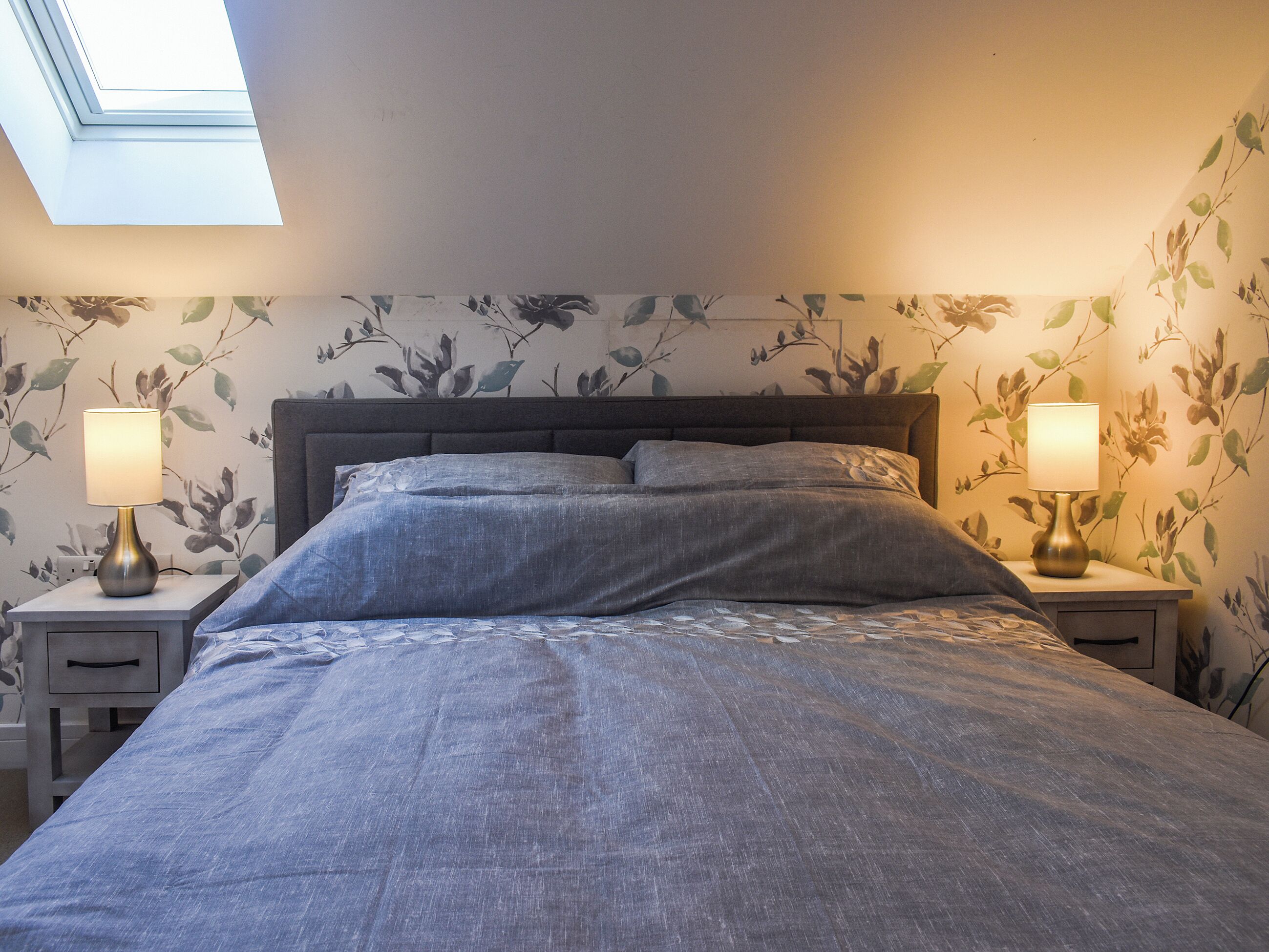 A bedroom with a skylight, a bed with grey bedding, two bedside lamps, and floral wallpaper.