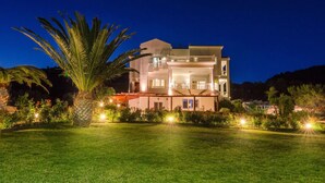 A two-story house with a balcony, surrounded by a well-lit lawn and palm trees. - A seafront villa with swimming pool (Achlia)