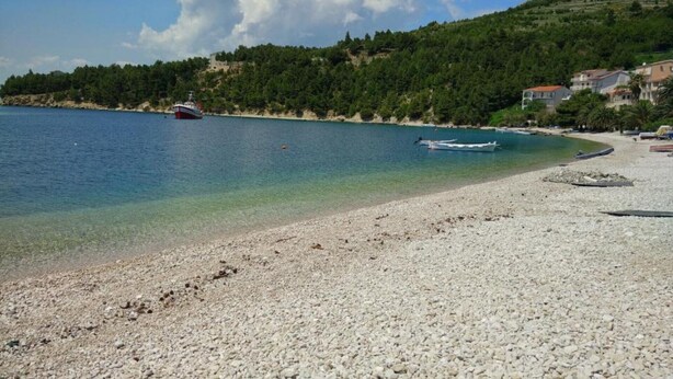 Una spiaggia di ciottoli con barche, un mare cristallino e una collina ricoperta di alberi sullo sfondo.