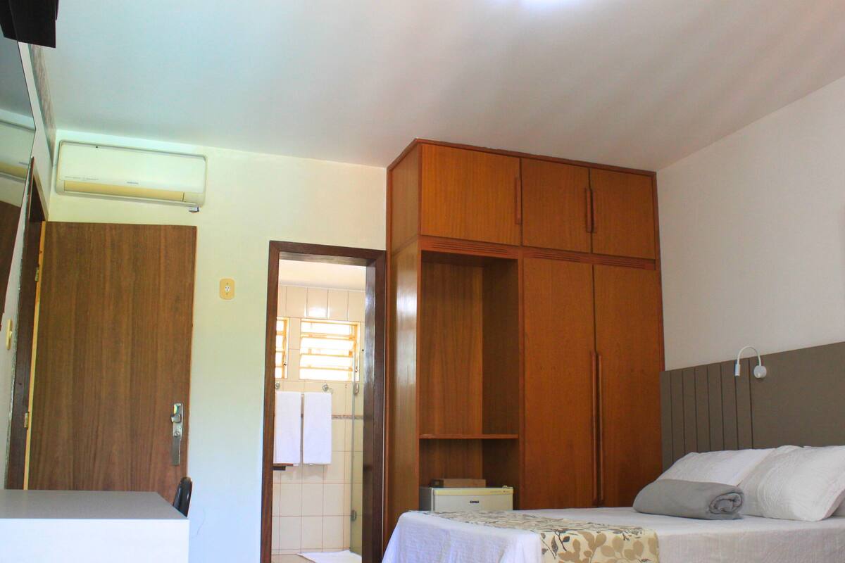 Habitación de hotel con cama, armario de madera, mesita de noche y baño visible a través de una puerta abierta.
