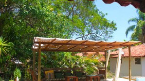 Un espacio de estar al aire libre cubierto con un banco y mesa de madera, rodeado de abundante vegetación y un cielo azul despejado.