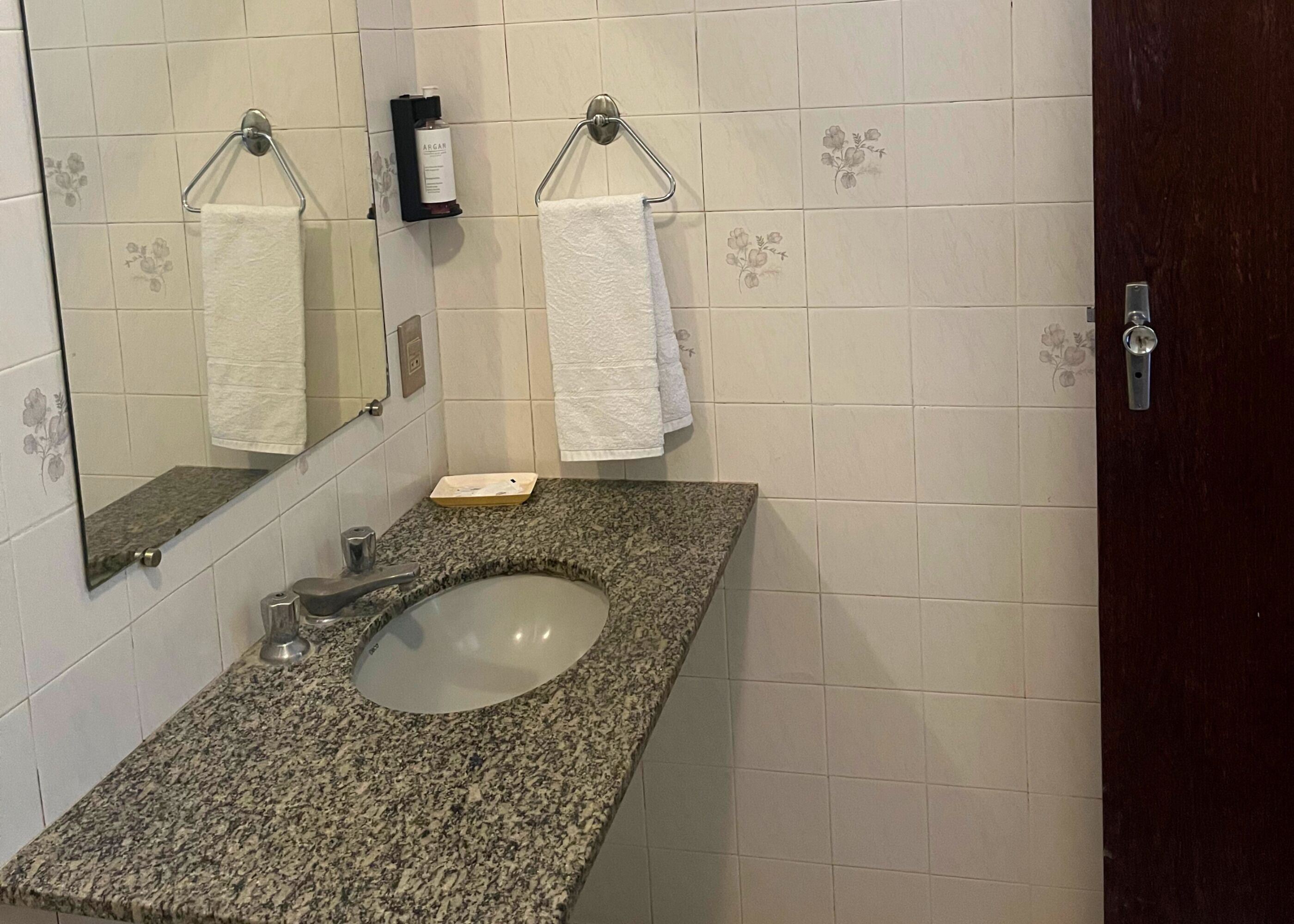 A bathroom with a granite countertop sink, two white towels hanging on towel racks, a mirror above the sink, and a soap dispenser on the wall.
