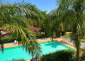 Une piscine entourée d’une végétation luxuriante et de palmiers, avec des chaises longues et une structure de toit carrelée en arrière-plan.